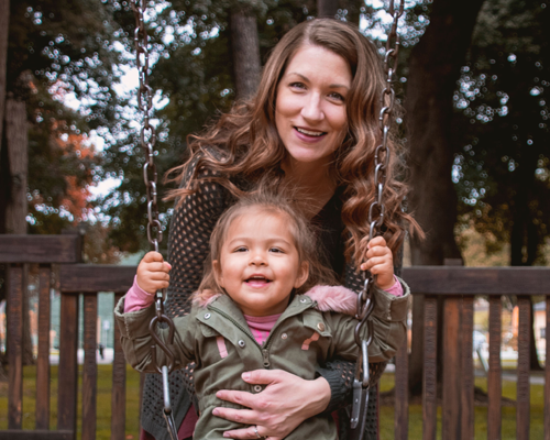 Michelle standing behind her young daughter and smiling. Her daughter is sitting on a swing and holding onto the chains.