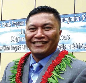 Headshot of Senator Kalani Kaneko from RMI. He is smiling and wearing a gray suit and two leis, one is red and one is green. 