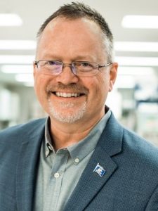 This is a headshot of Brent Sanford. He is wearing glasses, a gray shirt and gray suit jacket; the background appears to be a brightly lit office space.  He is smiling directly at the camera. 