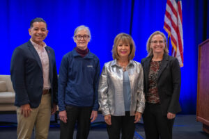 Four people stand in front of a stage, with a blue curtain behind them, next to a U.S. flag. There is one man and three women, all wearing business casual dress.