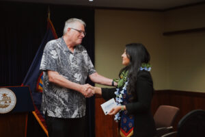 Dennis Mohatt stands left and shakes the hand of a recent graduate from the Guam Psychology Internship Program. The graduate is wearing a lei and holds her diploma certificate in one hand while shaking Dennis' hand.